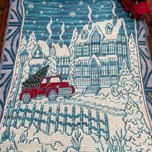 May include: A crocheted blanket with a winter scene featuring a house, a red truck with a Christmas tree in the back, and a white picket fence. The blanket is blue and white with a red border.
