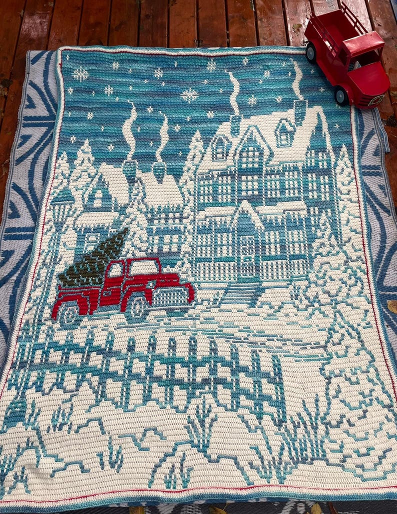 May include: A crocheted blanket with a winter scene featuring a house, a red truck with a Christmas tree in the back, and a white picket fence. The blanket is blue and white with a red border.