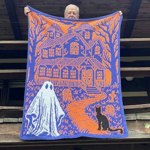 Op de afbeelding: Een gebreide Halloween-deken met een spookhuis, een geest en een zwarte kat in oranje en wit op een blauwe achtergrond. De deken wordt getoond, waarbij het gedetailleerde ontwerp wordt benadrukt.