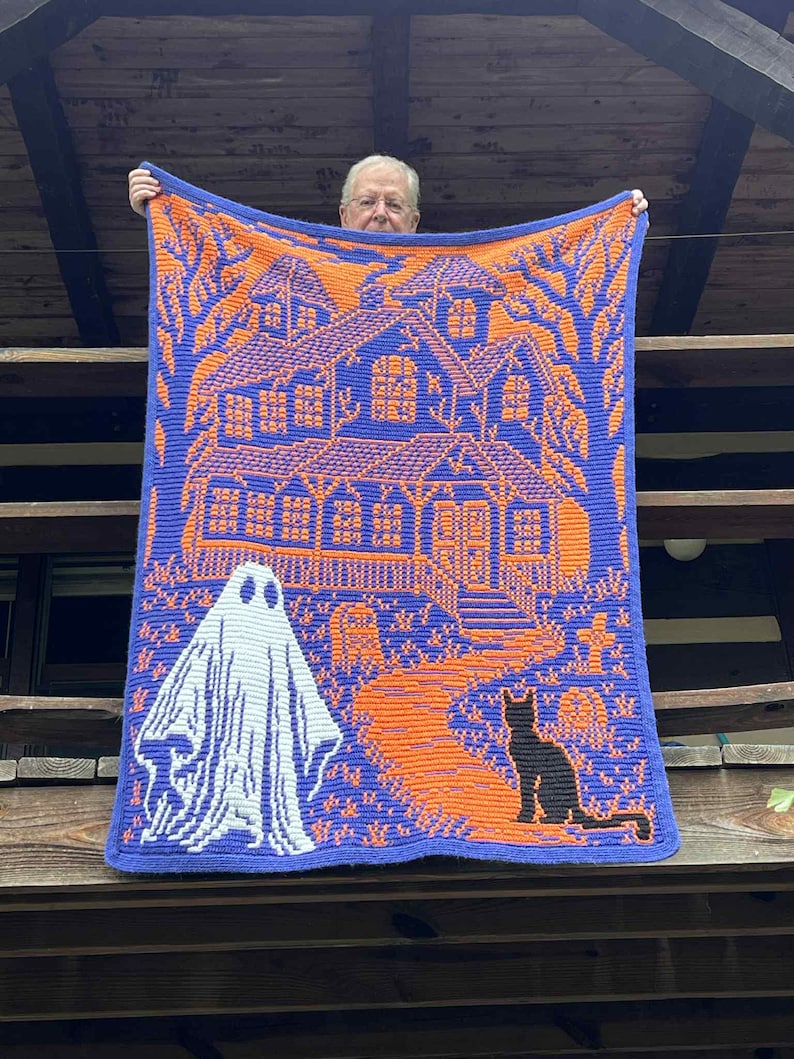 Op de afbeelding: Een gebreide Halloween-deken met een spookhuis, een geest en een zwarte kat in oranje en wit op een blauwe achtergrond. De deken wordt getoond, waarbij het gedetailleerde ontwerp wordt benadrukt.