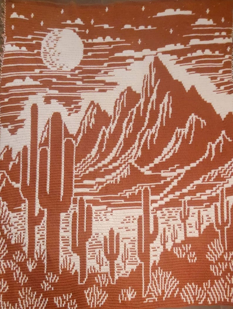 Peut inclure: Une couverture au crochet avec un motif de paysage d&eacute;sertique. La couverture repr&eacute;sente une cha&icirc;ne de montagnes, des cactus et une pleine lune dans un ciel &eacute;toil&eacute;. Les couleurs sont le brun et le blanc.