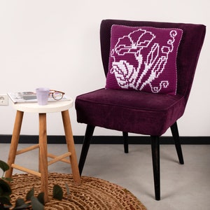 May include: A purple velvet armchair with a white and purple crocheted pillow featuring a floral design. The chair is sitting on a brown jute rug.