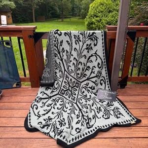 May include: A rectangular crochet blanket featuring a high-contrast black and light gray floral damask pattern. The design includes a central diamond motif surrounded by scrolling foliage and a solid black border. A small tag reads Handmade By Paula.