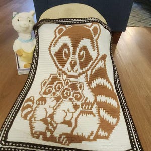May include: A crocheted blanket with a brown and white raccoon family design. The blanket has a white background with a brown raccoon and two smaller raccoons. The border is brown with a decorative stitch.