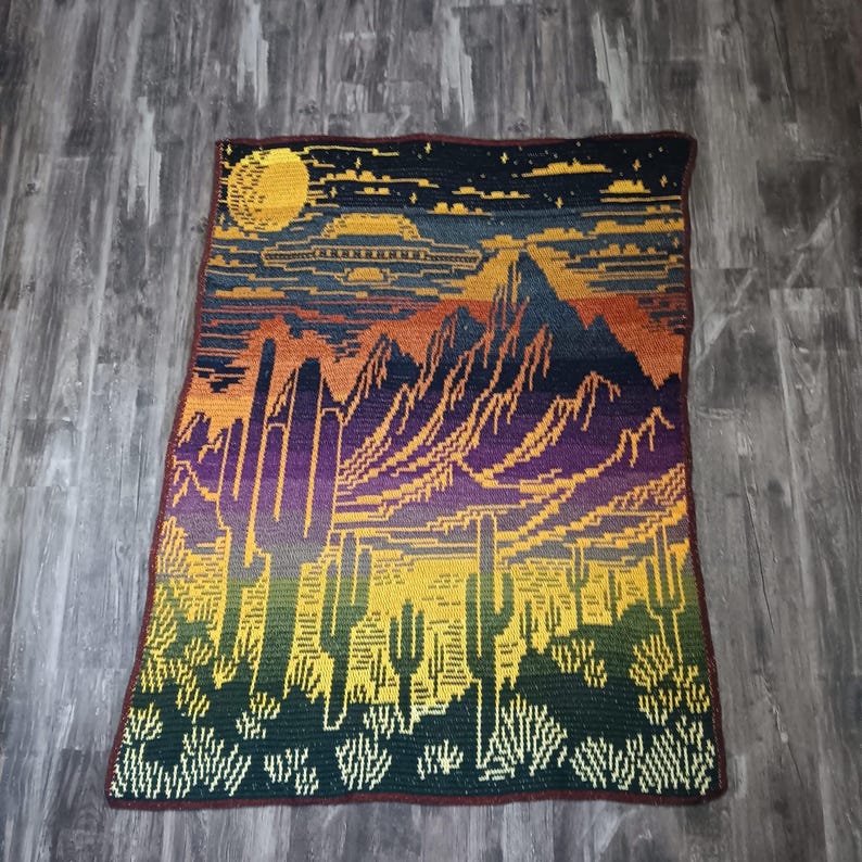 Peut inclure: Une couverture au crochet avec un motif de paysage d&eacute;sertique. La couverture repr&eacute;sente un ciel de coucher de soleil avec un OVNI, des montagnes et des cactus. Les couleurs sont jaune, orange, violet, vert et marron.