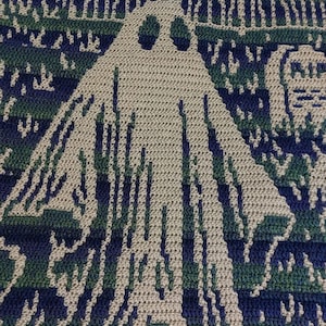 Op de afbeelding: Gehaakte deken met een spookontwerp in beige, groen en marineblauw. De deken toont een spookachtige sc&egrave;ne met een spook, een huis en een begraafplaats. Het haakwerk is gedetailleerd en cre&euml;ert een textuureffect.