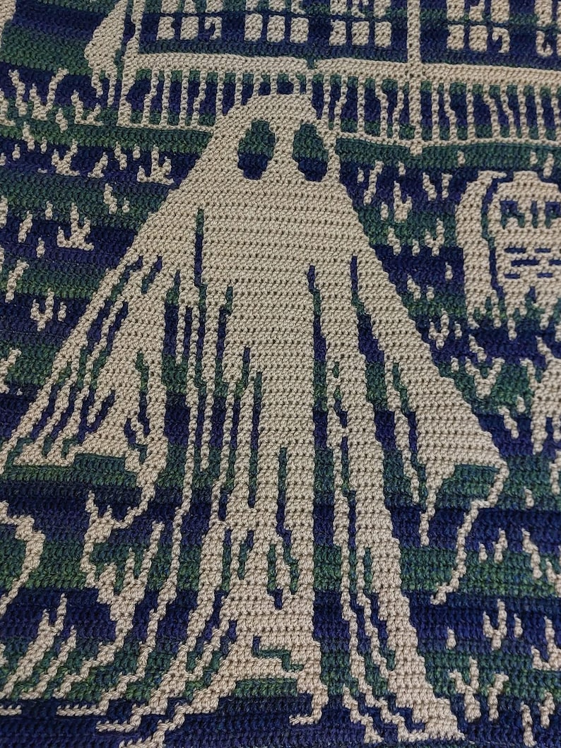 Op de afbeelding: Gehaakte deken met een spookontwerp in beige, groen en marineblauw. De deken toont een spookachtige sc&egrave;ne met een spook, een huis en een begraafplaats. Het haakwerk is gedetailleerd en cre&euml;ert een textuureffect.