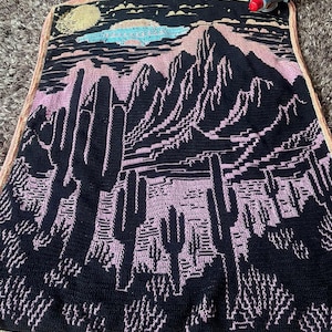 Peut inclure: Une couverture en crochet noire et rose avec un motif de paysage d&eacute;sertique. La couverture repr&eacute;sente des montagnes, des cactus et une lune dans le ciel.