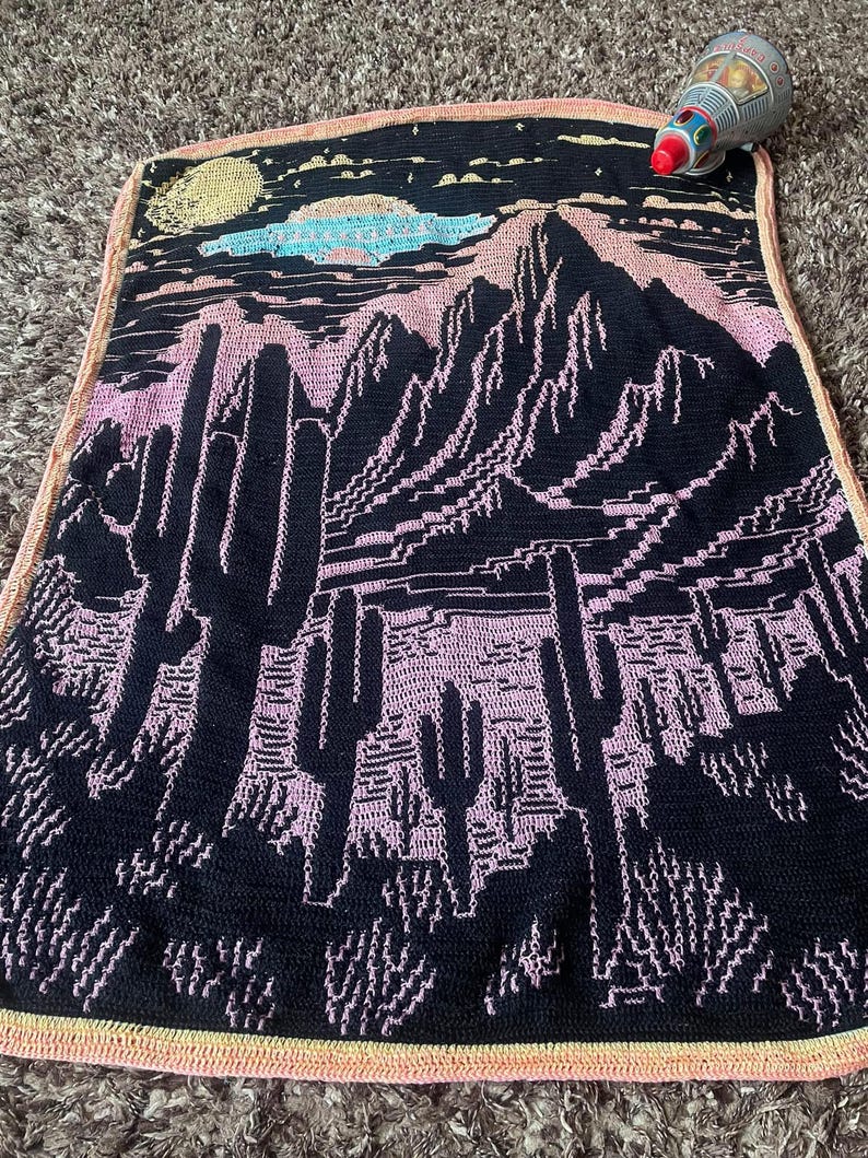 Peut inclure: Une couverture en crochet noire et rose avec un motif de paysage d&eacute;sertique. La couverture repr&eacute;sente des montagnes, des cactus et une lune dans le ciel.