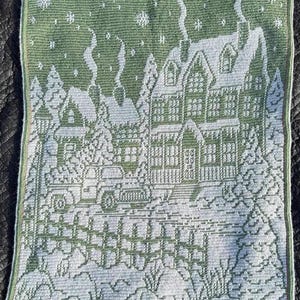 May include: A green and white crocheted wall hanging depicting a winter scene with houses, trees, and snow. The houses have white roofs and green windows. The trees are green and the snow is white.