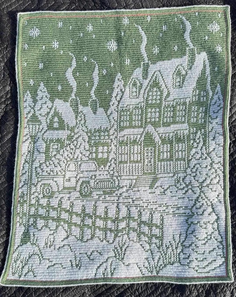May include: A green and white crocheted wall hanging depicting a winter scene with houses, trees, and snow. The houses have white roofs and green windows. The trees are green and the snow is white.