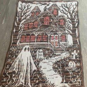 Op de afbeelding: Een gehaakte deken met een spookhuis sc&egrave;ne. De deken toont een bruin huis met roze ramen, een witte geest en een begraafplaats. De deken heeft een bruin-witte rand, perfect voor Halloween.