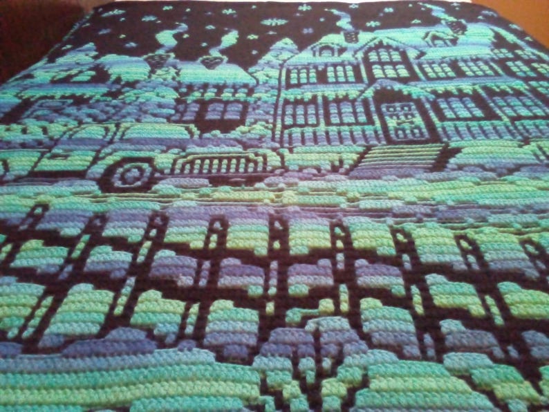 May include: A crocheted blanket with a winter scene featuring a house, a car, and a fence. The blanket is made with blue, green, and purple yarn.