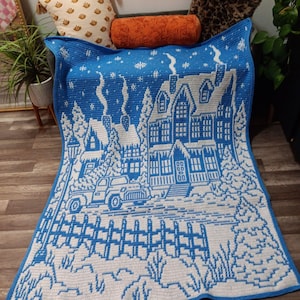 May include: A blue and white crocheted blanket with a winter scene of a house, trees, a truck, and a fence. The blanket is on a wooden floor.