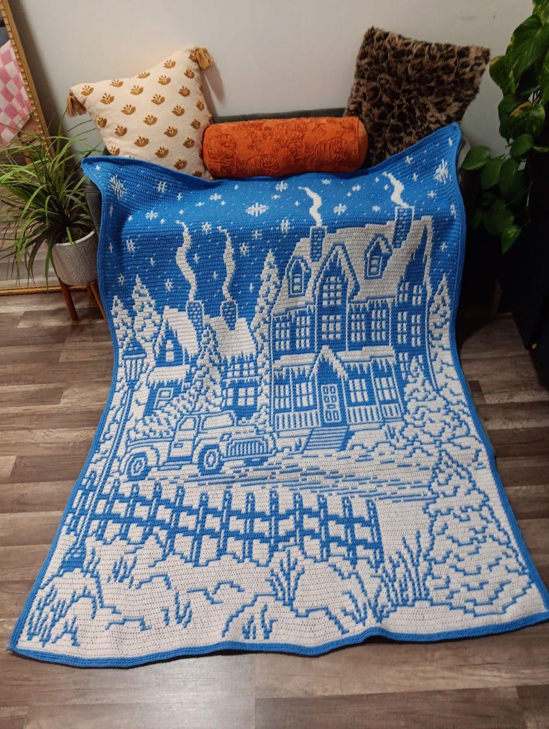 May include: A blue and white crocheted blanket with a winter scene of a house, trees, a truck, and a fence. The blanket is on a wooden floor.