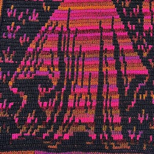 Op de afbeelding: Een handgemaakte gehaakte deken met een geometrisch ontwerp in zwart, roze, oranje en paars. De deken heeft een herhalend patroon van een gestileerde figuur. De randen zijn afgewerkt met een roze en paarse rand.