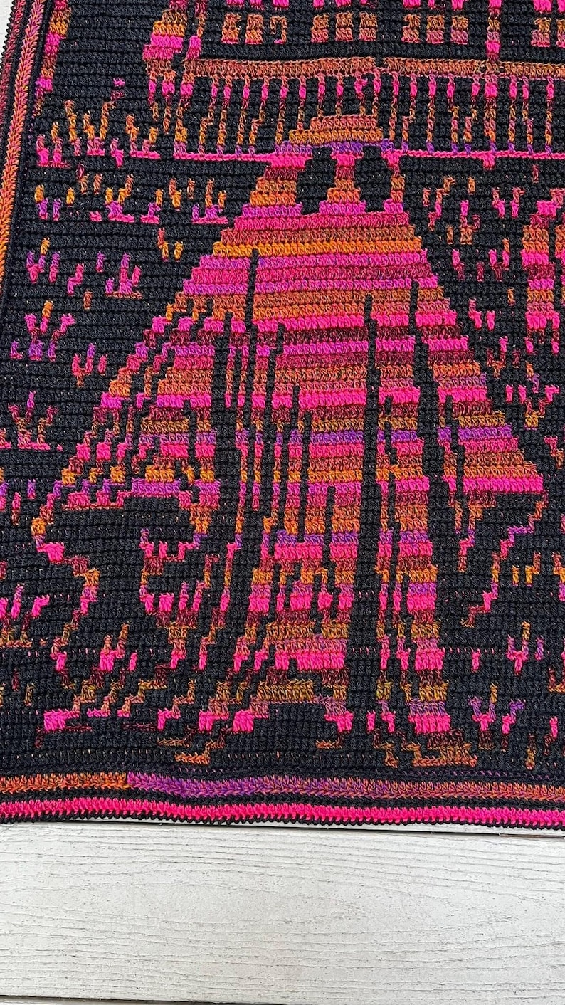 Op de afbeelding: Een handgemaakte gehaakte deken met een geometrisch ontwerp in zwart, roze, oranje en paars. De deken heeft een herhalend patroon van een gestileerde figuur. De randen zijn afgewerkt met een roze en paarse rand.