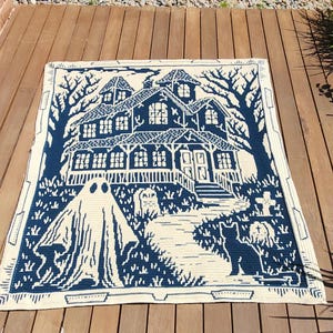 Op de afbeelding: Een cr&egrave;me en marineblauwe gehaakte deken met een spookhuissc&egrave;ne. Het ontwerp omvat een geest, kat, grafstenen en bomen. De deken heeft een rechthoekige vorm en een decoratieve rand.