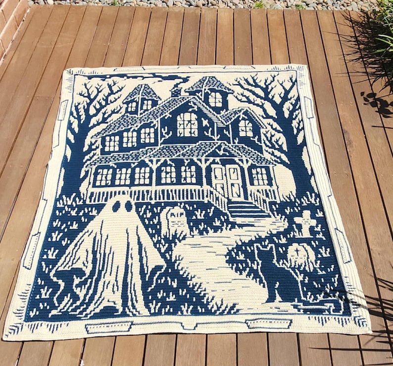 Op de afbeelding: Een cr&egrave;me en marineblauwe gehaakte deken met een spookhuissc&egrave;ne. Het ontwerp omvat een geest, kat, grafstenen en bomen. De deken heeft een rechthoekige vorm en een decoratieve rand.