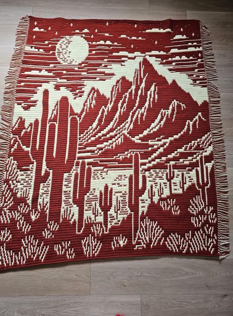 Peut inclure: Une couverture en crochet repr&eacute;sentant une sc&egrave;ne de d&eacute;sert avec des montagnes, des cactus et une pleine lune. La couverture est r&eacute;alis&eacute;e en fil rouge et blanc et poss&egrave;de une bordure en franges.