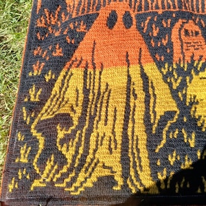 Op de afbeelding: Een Halloween-deken met een spook, een spookhuis en een kerkhof in oranje, geel en zwart. De deken is gemaakt van een textuurrijk materiaal, mogelijk gebreid of gehaakt. Het ontwerp is een herhalend patroon.