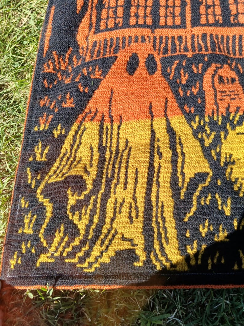 Op de afbeelding: Een Halloween-deken met een spook, een spookhuis en een kerkhof in oranje, geel en zwart. De deken is gemaakt van een textuurrijk materiaal, mogelijk gebreid of gehaakt. Het ontwerp is een herhalend patroon.
