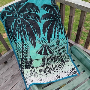 Peut inclure: Une couverture de plage en crochet avec un dégradé de bleu, vert et blanc. La couverture présente un motif de palmiers et un parasol.