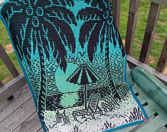 Peut inclure: Une couverture de plage en crochet avec un dégradé de bleu, vert et blanc. La couverture présente un motif de palmiers et un parasol.