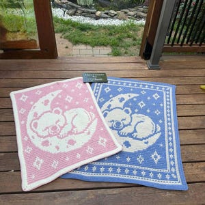 May include: Two handmade baby blankets. One is pink with a white bear on a crescent moon and stars design. The other is blue with a white bear on a crescent moon and stars design. Both have a white border.