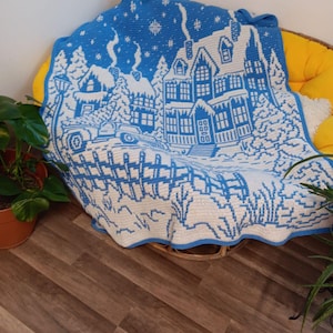 May include: A blue and white crocheted blanket with a winter scene of a house, trees, and a car. The blanket is on a wicker chair.