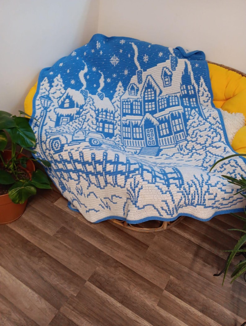 May include: A blue and white crocheted blanket with a winter scene of a house, trees, and a car. The blanket is on a wicker chair.