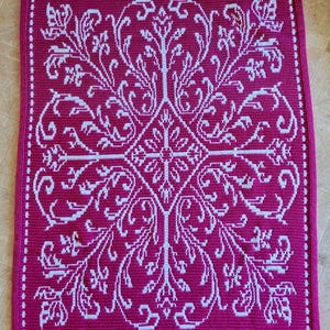 Pode incluir: Um cobertor de crochê magenta com um design floral e geométrico branco. O cobertor apresenta uma forma de diamante central com padrões florais radiantes e uma borda pontilhada. O design é simétrico e detalhado, adequado para decoração de casa.