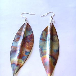 May include: Two leaf-shaped earrings with a metallic finish. The earrings are teardrop-shaped and hang from silver-coloured ear wires. The surface of the earrings has a blend of colours, including bronze, blue, and gold.