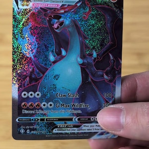 Puede incluir: Una carta Pokémon Charizard VMAX con un Charizard Gigantamax. La carta tiene un fondo holográfico con una combinación de colores azul y rojo. El texto de la carta incluye "Claw Slash" y "G-Max Wildfire".