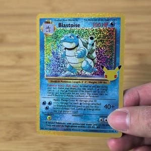 Peut inclure: Carte Pokémon holographique Blastoise. La carte présente une illustration bleue de Blastoise avec une carapace et des jets d'eau. La carte porte les mentions "Blastoise" et "100 HP". La carte mesure 1,60 m de long et pèse 85,7 kg.