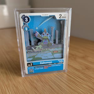 May include: A 3D layered shadow box art piece featuring the Digimon Gabumon card. The card displays the character Gabumon in a blue and white setting with text including Gabumon, Lv.3, and 2000 DP, all encased in a clear protective plastic display case.