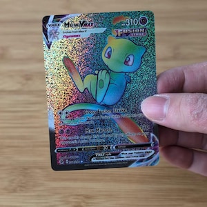 Puede incluir: Una tarjeta Pokémon VMAX Mew VMAX Fusion Strike holográfica. La tarjeta presenta una criatura de dibujos animados verde y rosa con el texto "Cross Fusion Strike" y "Max Miracle". La tarjeta tiene 310 PS.