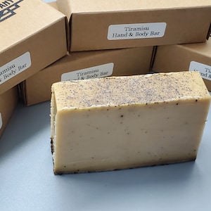 May include: A close-up of a light brown bar of soap with a light brown and black speckled top. The soap is labeled "Tiramisu Hand & Body Bar".