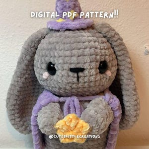 Puede incluir: Un conejito de crochet gris que lleva un sombrero morado con una estrella en la parte superior. El conejito sostiene una estrella amarilla en sus patas. El texto "DIGITAL PDF PATTERN!!" está escrito encima del conejito. El texto "@cuteelittlecreations" está escrito debajo del conejito.
