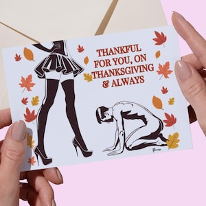 May include: A greeting card with the text "THANKFUL FOR YOU, ON THANKSGIVING & ALWAYS" and illustrations of a woman in a skirt and high heels, and a person kneeling. The card is held in front of a cream-colored envelope, with fall leaves.