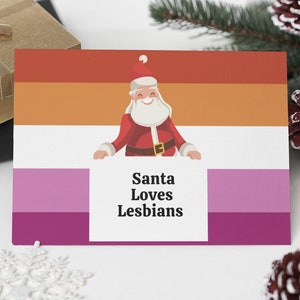 May include: A greeting card with the text "Santa Loves Lesbians" and a Santa Claus illustration. The card features the lesbian pride flag colors. The image is surrounded by Christmas decorations, including pine cones and gift boxes.