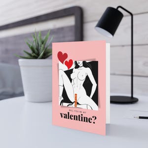 May include: A pink Valentine's Day card with a black and white illustration of a woman and two red hearts. The card reads "WILL YOU BE MY VALENTINE?" in black text.