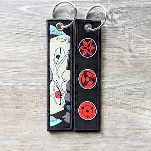 May include: Two black keychains with silver key rings. One keychain features a black and white anime character with a red eye. The other keychain has three red and black symbols.