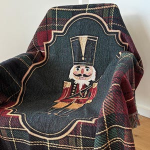Puede incluir: Una manta tejida con un diseño a cuadros en negro, verde y rojo, colocada sobre una silla. El centro presenta una ilustración detallada de un soldado cascanueces en un marco decorativo. La manta tiene un borde con flecos.