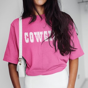 May include: A magenta t-shirt with the word "COWGIRL" in white lettering, worn with a white skirt and a white crossbody bag. The outfit is completed with dark nail polish.