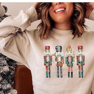 May include: Beige sweatshirt featuring a watercolor illustration of four toy nutcrackers. The nutcrackers are in various colors, including red, green, and gold, and are standing in a row. The sweatshirt is a neutral color, and the overall theme is holiday-themed.