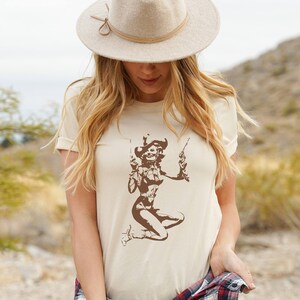 以下が含まれることがあります： ベージュのTシャツに、カウボーイハットをかぶり、2丁のピストルを持った女性の茶色のグラフィックがプリントされています。女性は短いスカートとブーツを履いています。Tシャツは、長いブロンドの髪とベージュのカウボーイハットをかぶった人が着用しています。