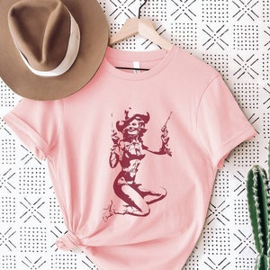 以下が含まれることがあります： ピンクのTシャツに、カウボーイハットをかぶり、2丁の拳銃を持った女性のシルエットがマロン色で描かれています。