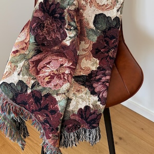 May include: A floral tapestry throw blanket draped over a brown leather chair. The blanket features a pattern of large burgundy, pink, and cream-colored flowers with green leaves. The blanket has a fringed edge with multiple colors.