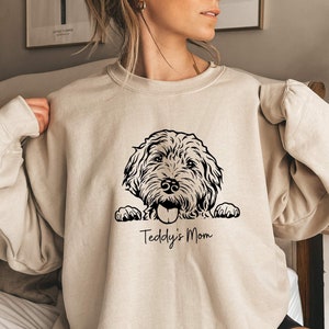 Może przedstawiać: Beżowa bluza z czarno-białą ilustracją psa wyglądającego zza górnej krawędzi. Pod psem nadrukowany jest tekst "Teddy's Mom".
